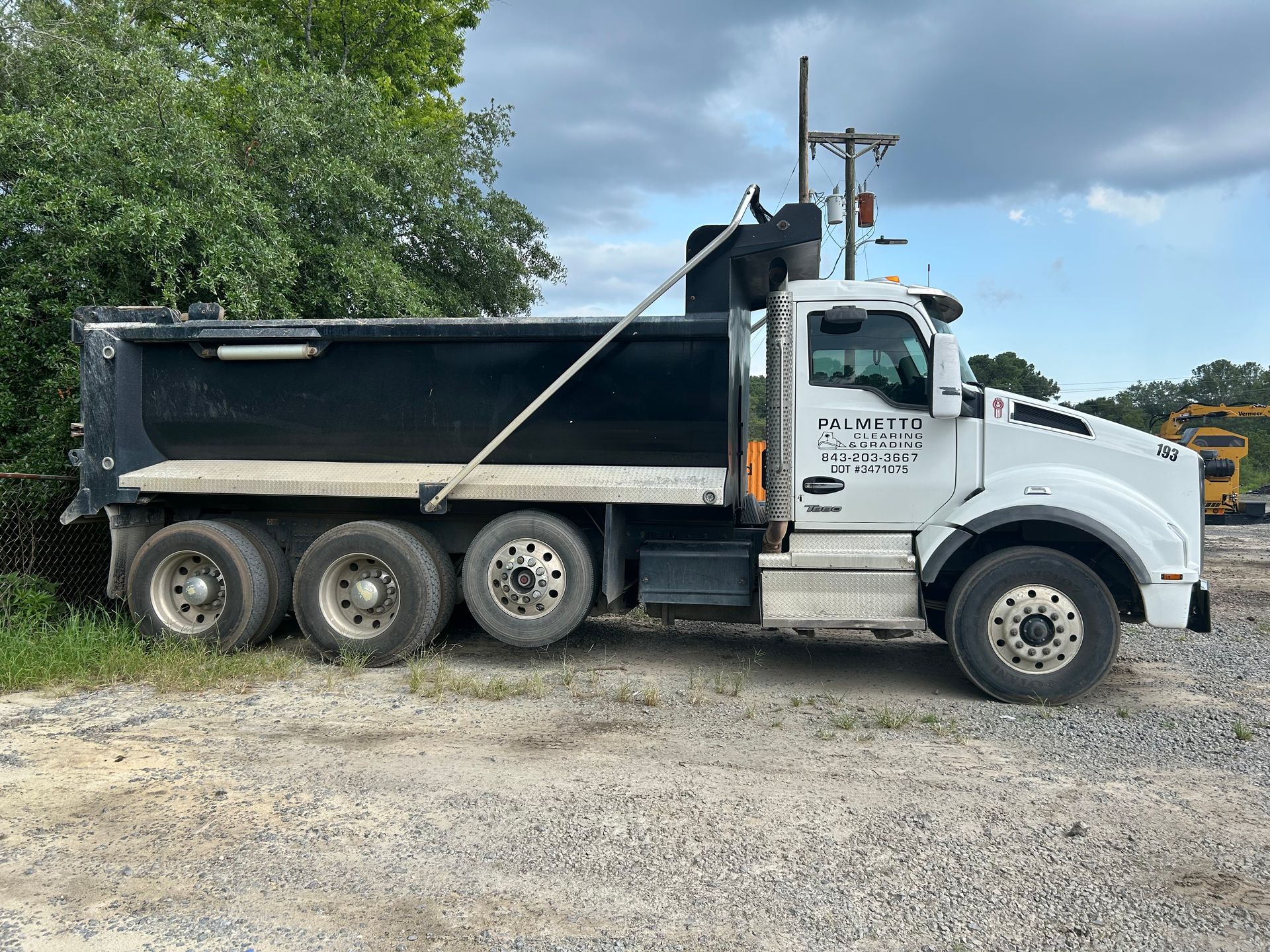A white dump truck with a black cargo bed parked on a gravel lot next to green trees under a cloudy sky.