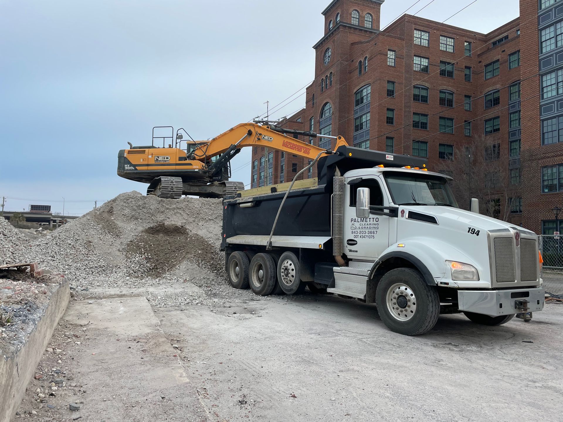 A yellow excavator loading a pile of rubble into a white dump truck parked in front of a brick building.