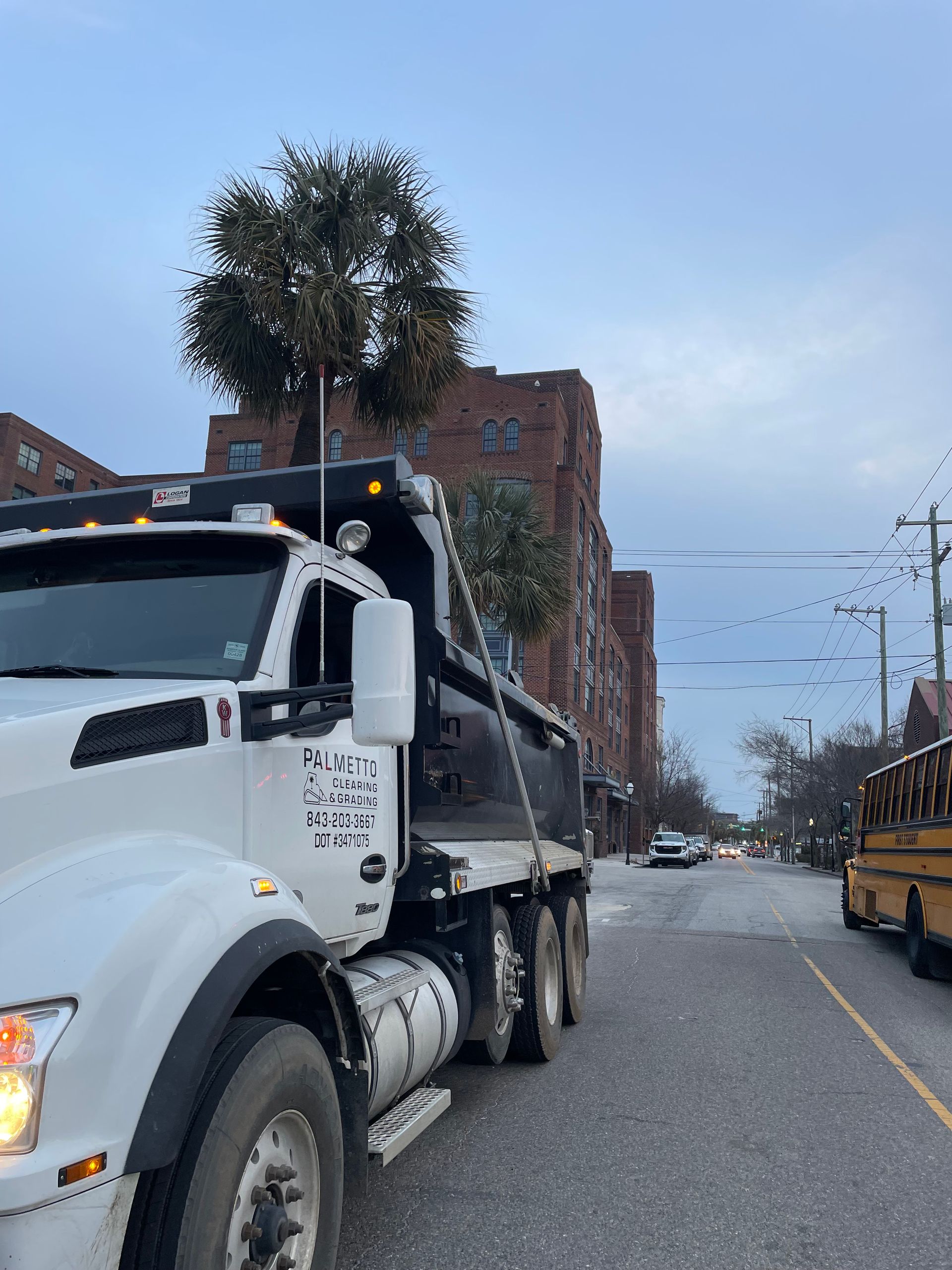 A white dump truck parked on a city street with tall palm trees nearby and a school bus visible in the distance.