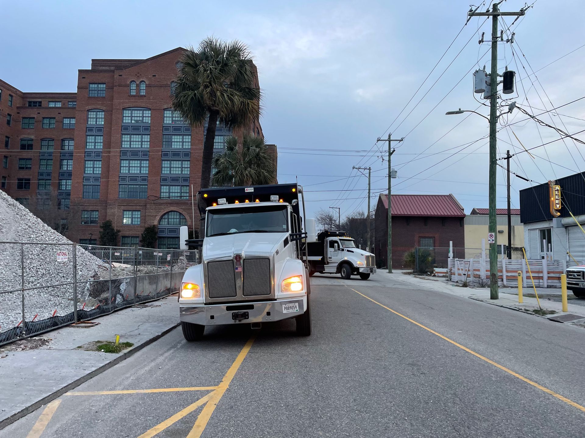 Two white dump trucks on an asphalt road next to a large pile of construction debris and a brick building.