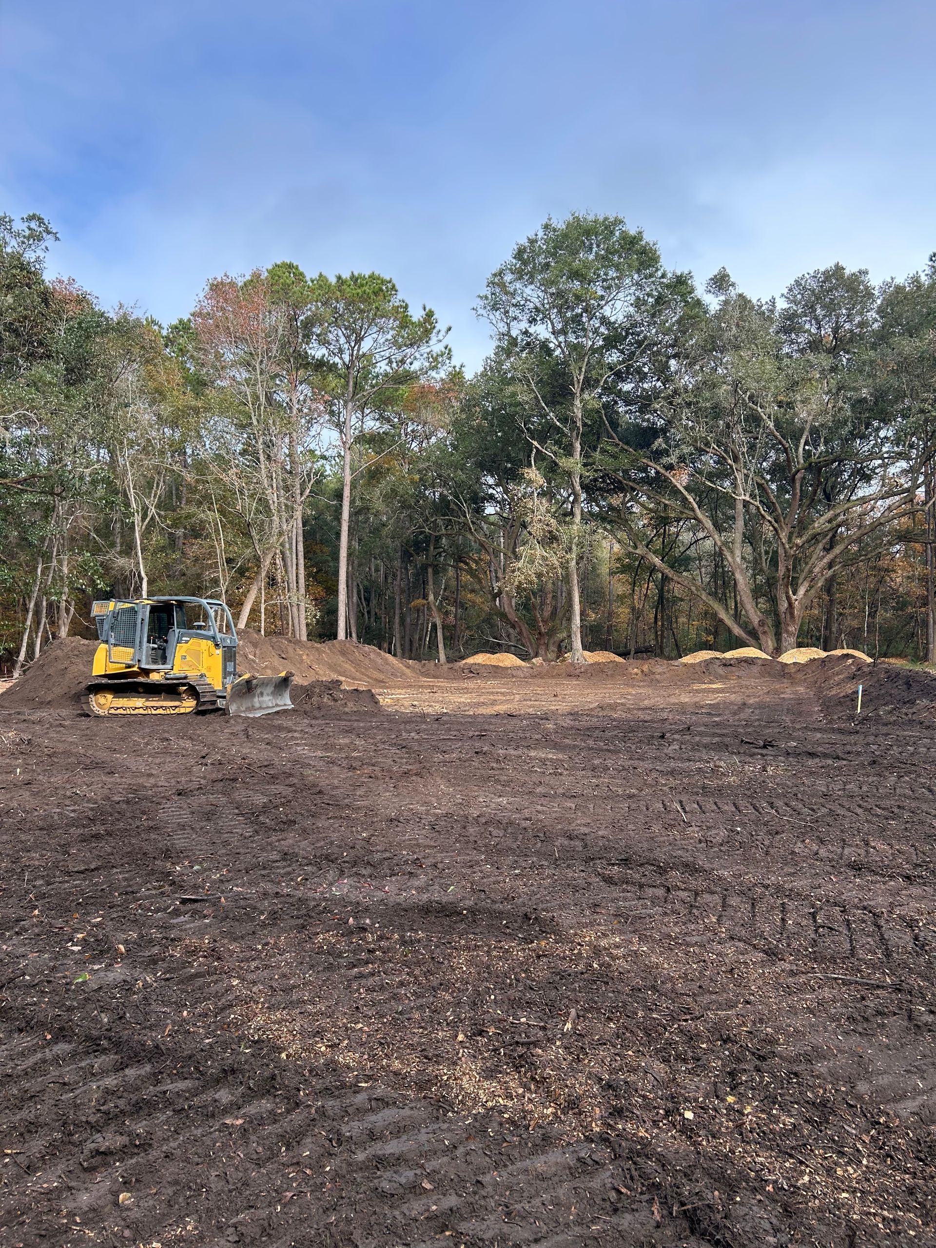 A yellow bulldozer clears dark soil on a construction site in front of a line of trees under a blue, cloudy sky.