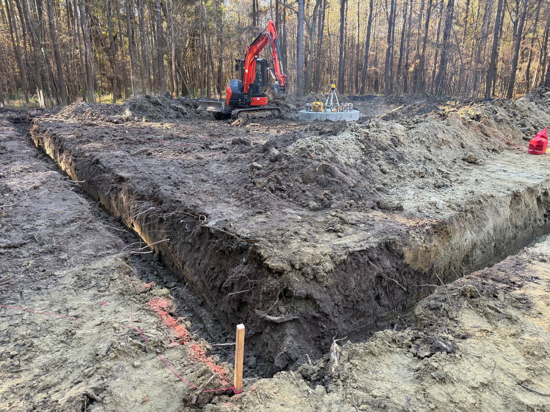 An orange excavator stands on a wooded lot next to a large rectangular foundation trench marked by orange string.