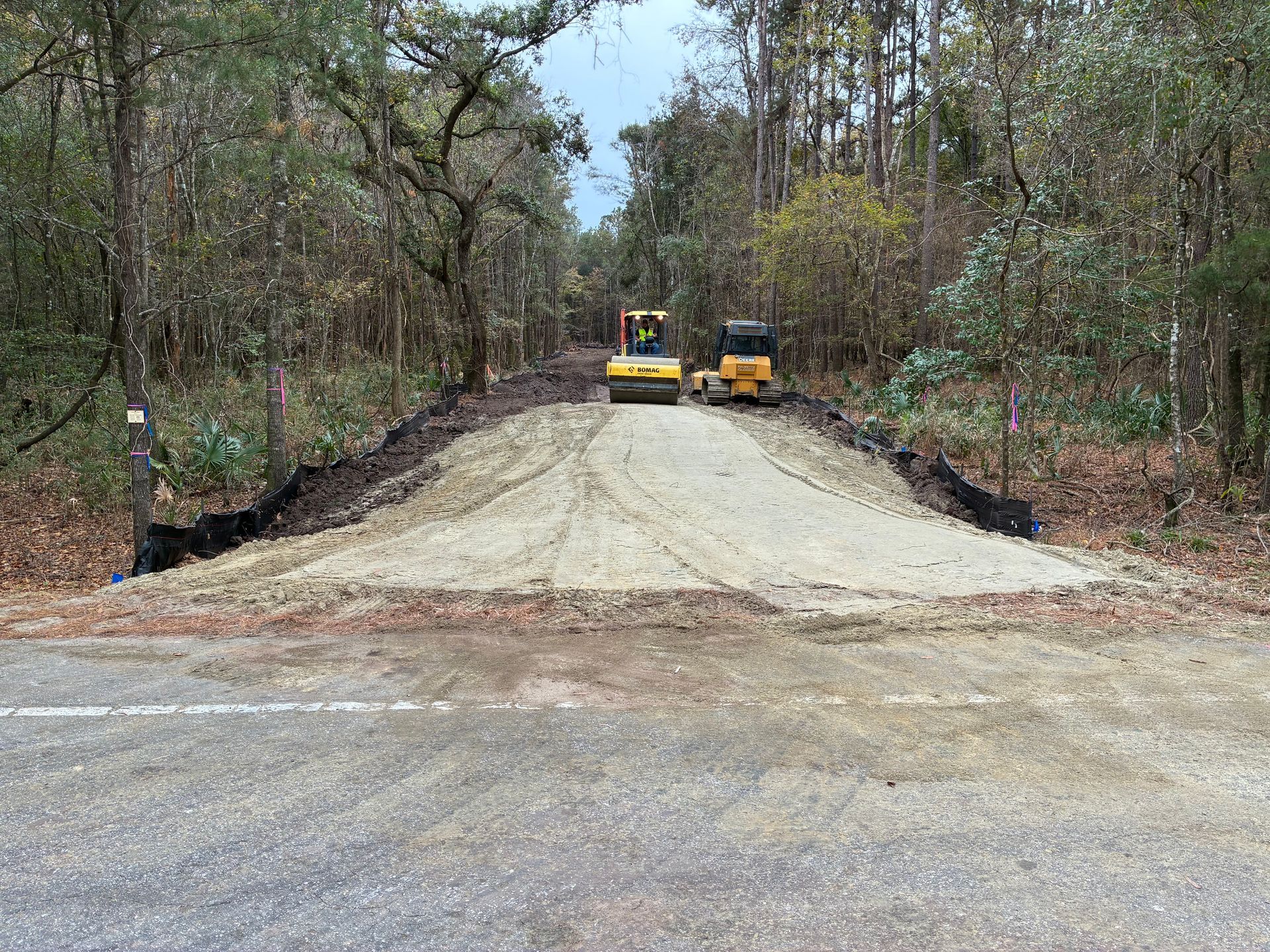 A road roller and a bulldozer work on a gravel driveway connecting to an asphalt road in a wooded area.