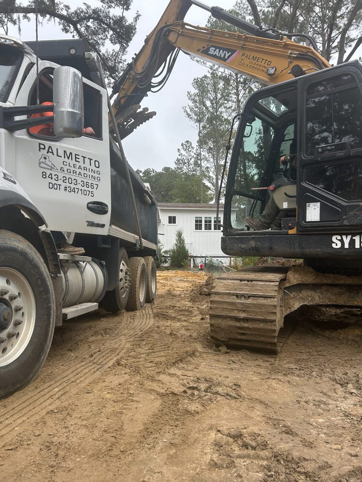 An excavator loading soil into a white Palmetto dump truck at a construction site.