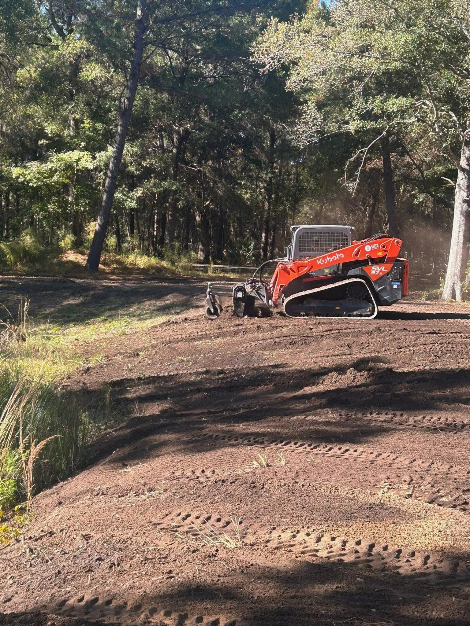 An orange track loader levels soil at the edge of a wooded area, leaving visible tread marks on the disturbed earth.