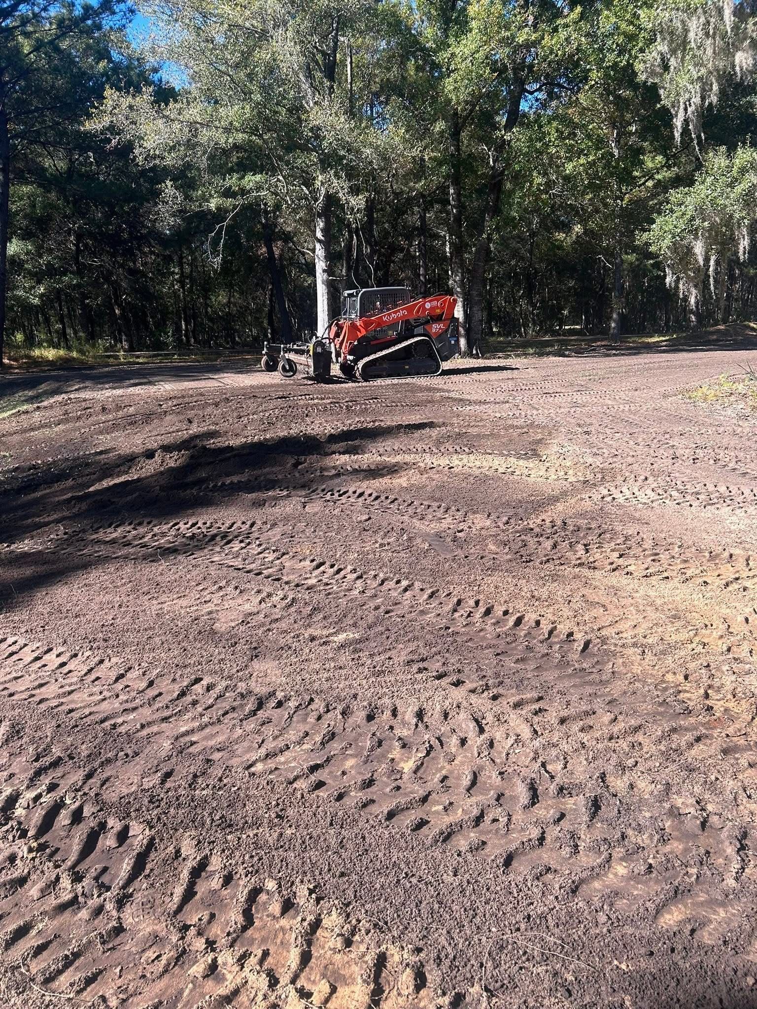 An orange tracked skid steer machine sits on a freshly cleared, muddy lot in front of a dense treeline.