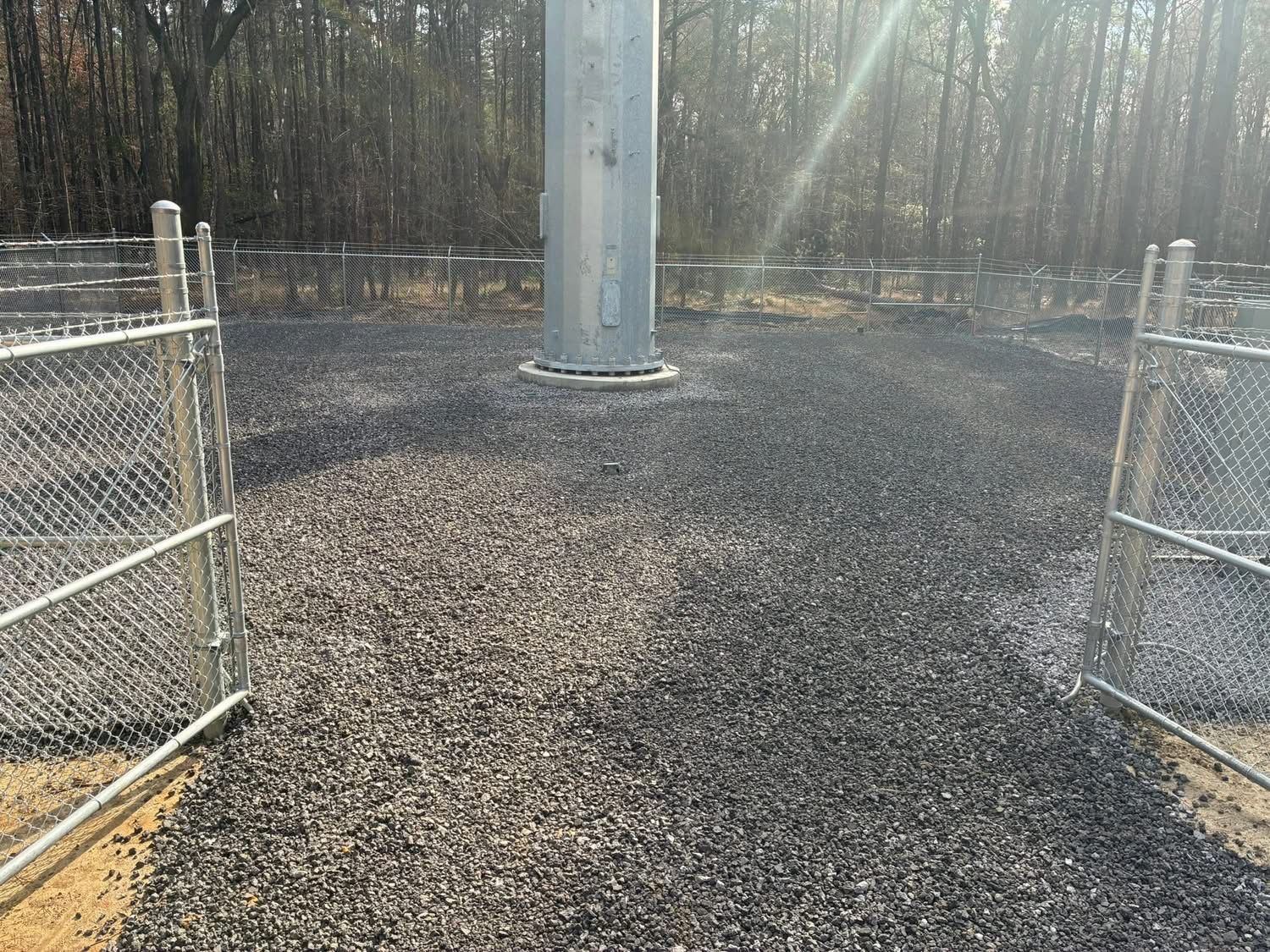 A metallic utility pole stands in the center of a graveled enclosure surrounded by a chain-link fence.