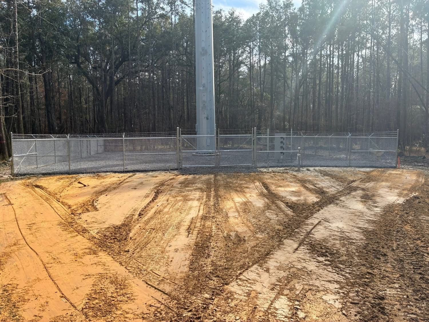 A metal pole rises from a dirt clearing enclosed by a chain-link fence, surrounded by a forest in the background.