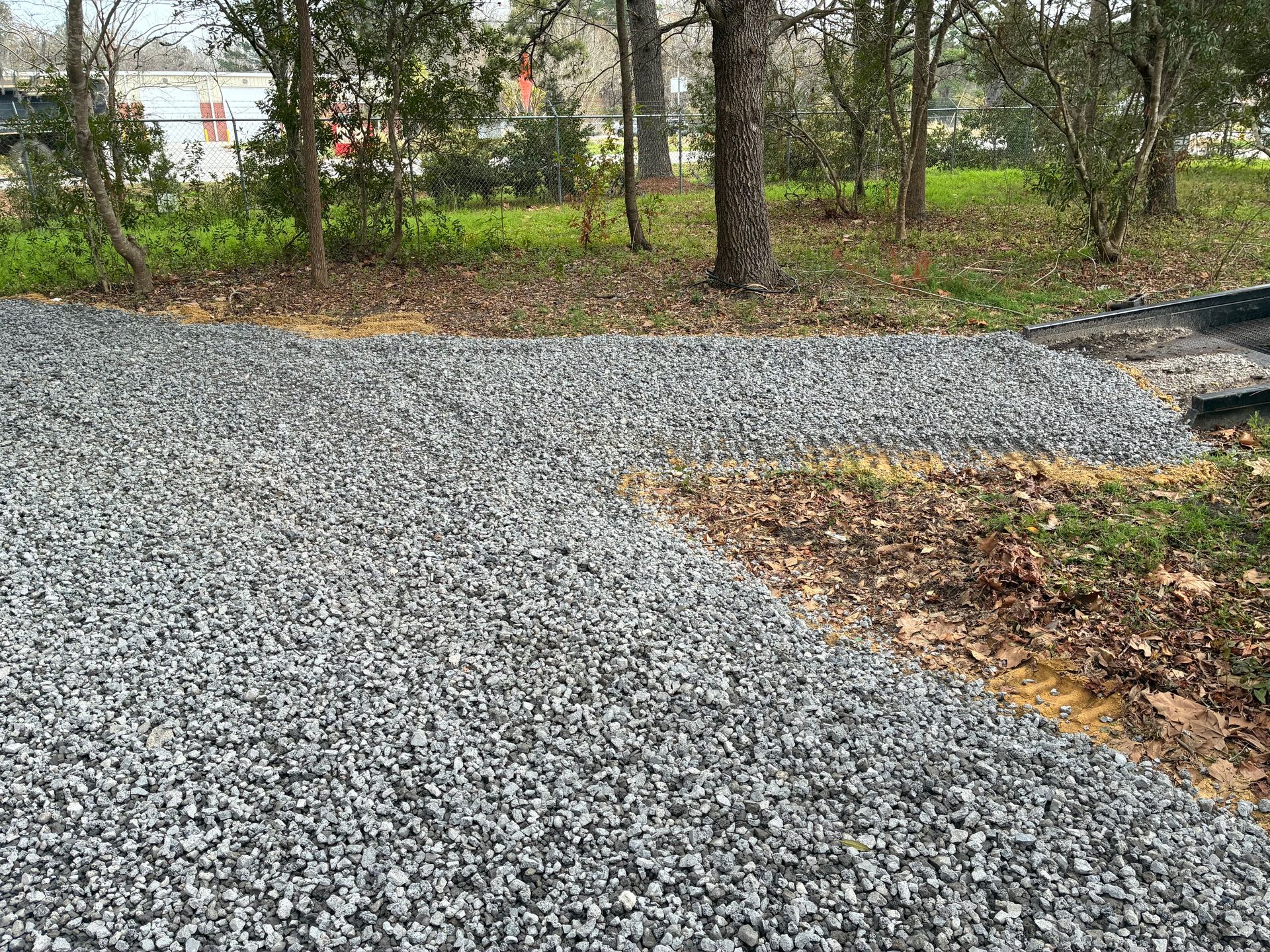 A gray gravel driveway transitions into a dirt pathway leading toward a wooded area with trees and bushes.