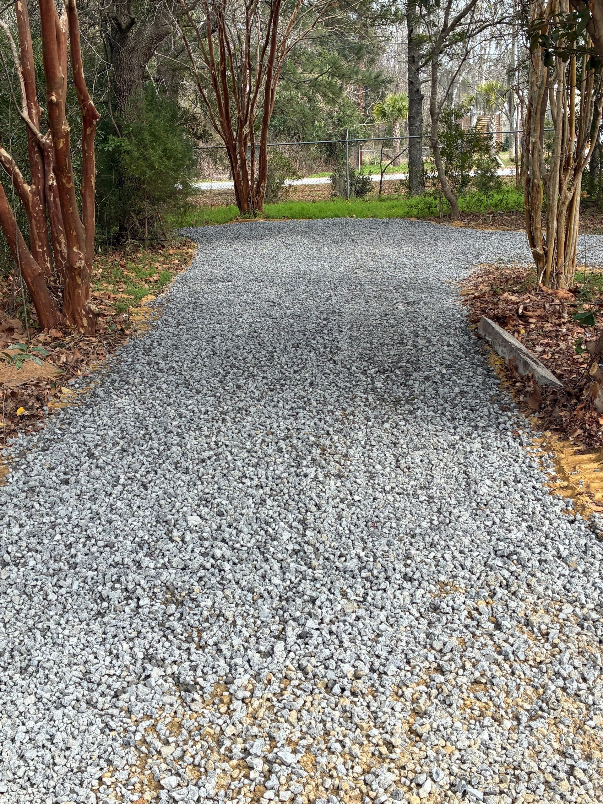 A gravel path leads through a yard with bare trees on both sides, transitioning from tan gravel to grey stones.