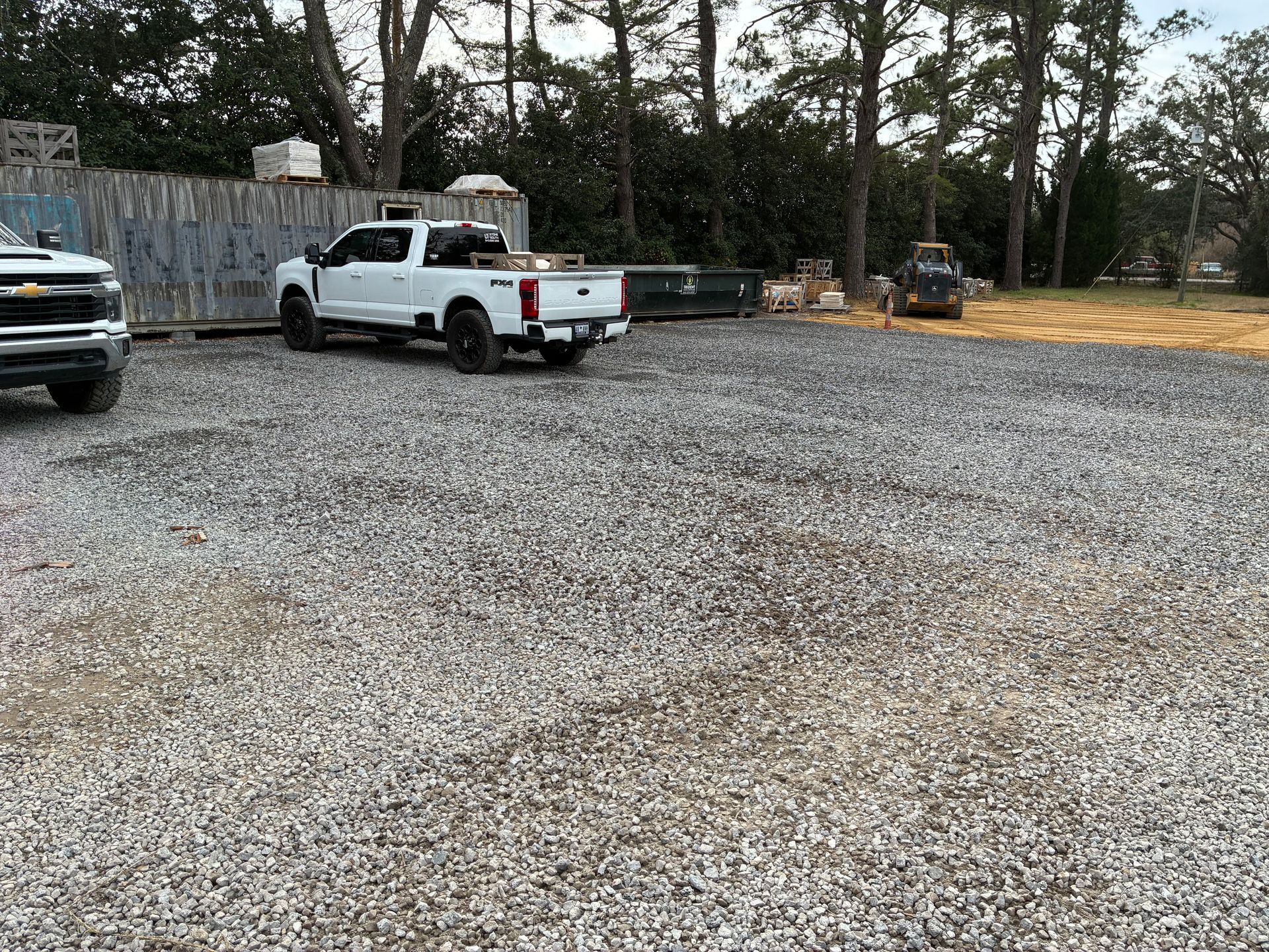 A white pickup truck parked on a gravel lot next to a shipping container, with a construction vehicle in the distance.