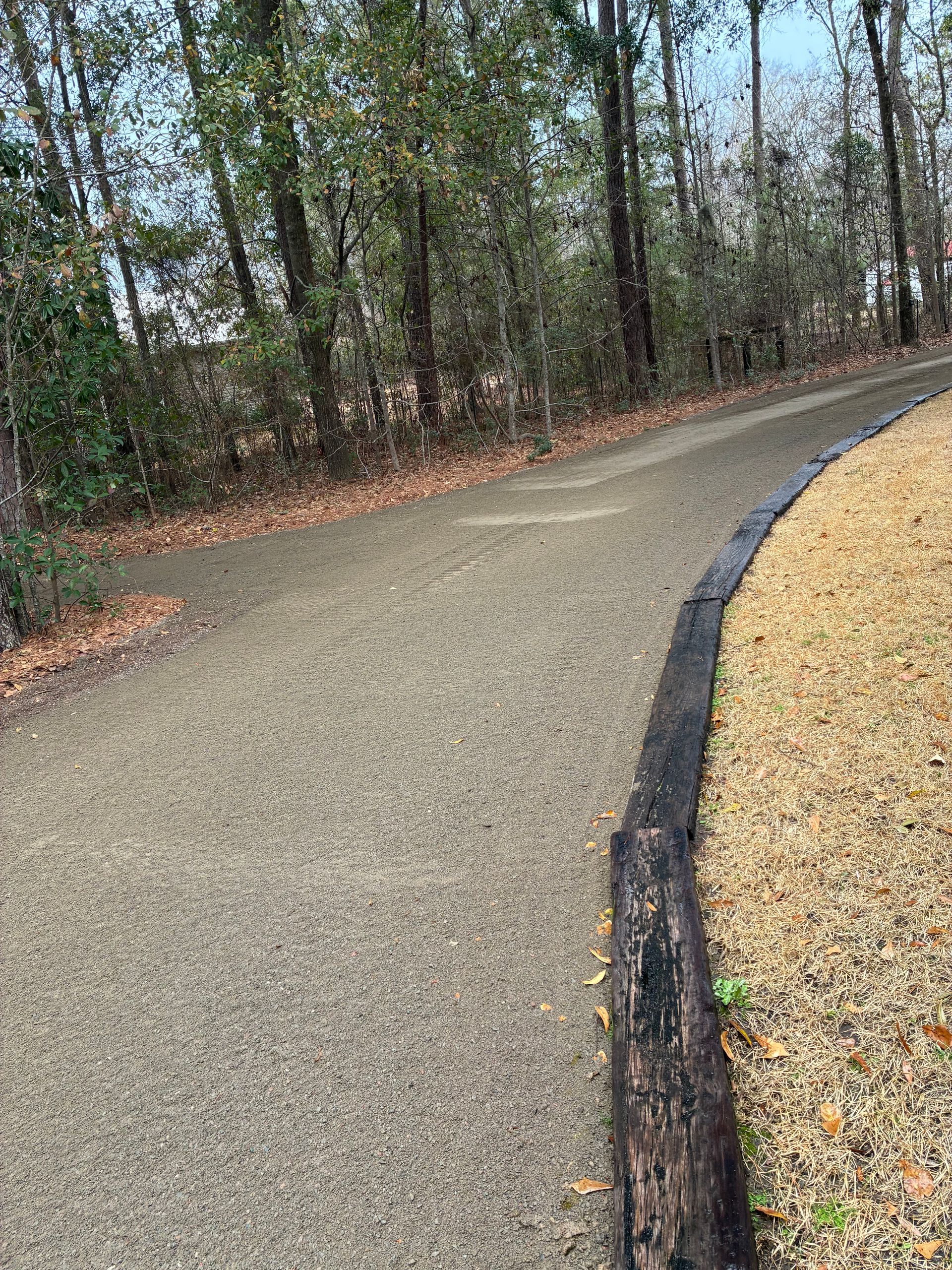 A gravel pathway curves through a wooded area, bordered by wooden beams on the right and surrounded by trees and leaf litter.
