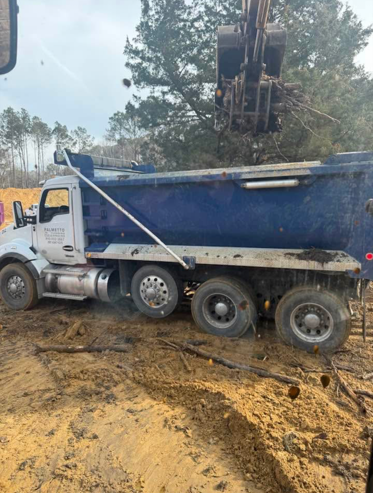A blue dump truck on a dirt construction site is being loaded with tree debris by an excavator.