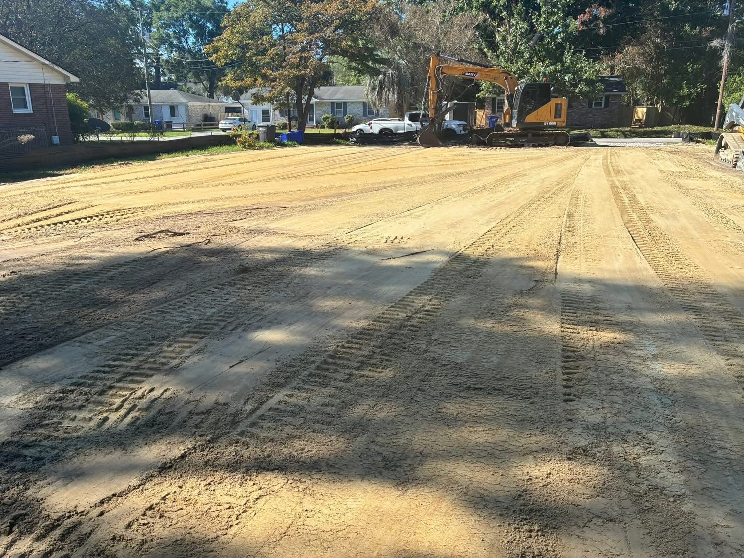 A construction site with tire tracks on a cleared, sandy lot and an excavator in the background near suburban houses.