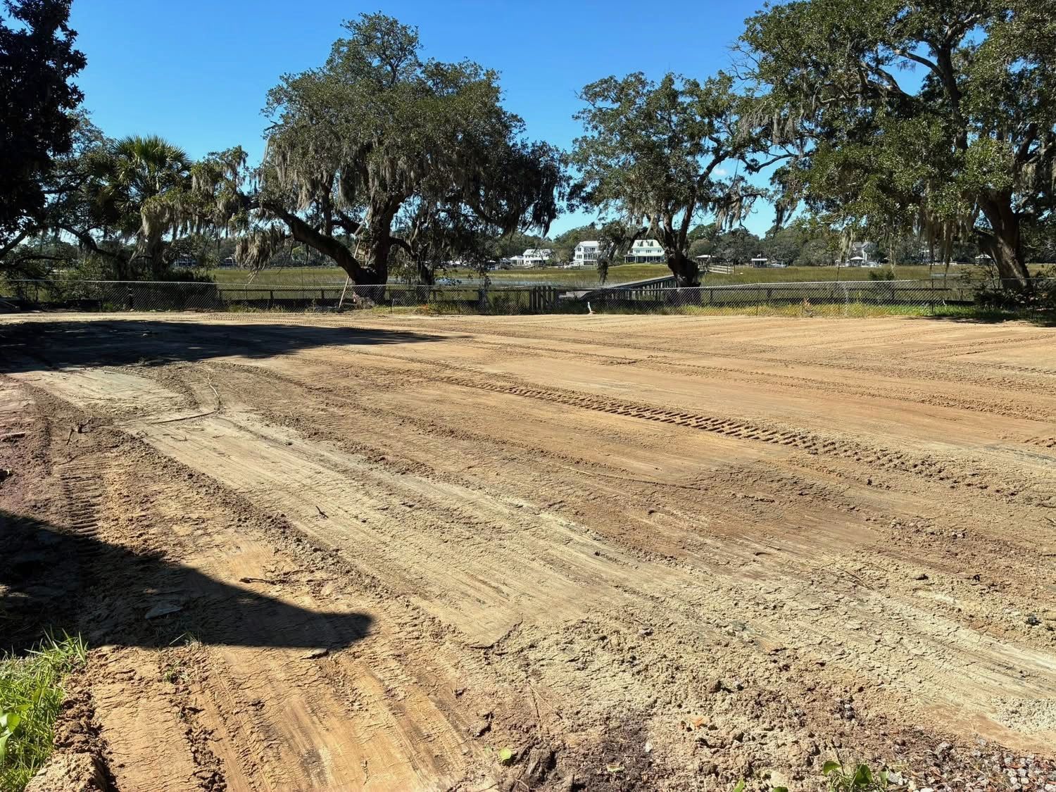 A cleared, level dirt lot with tire tracks, bordered by large oak trees under a clear blue sky.