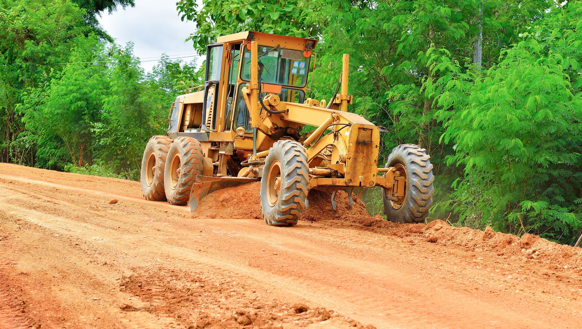 Yellow grader smoothing a red dirt road in a wooded area.