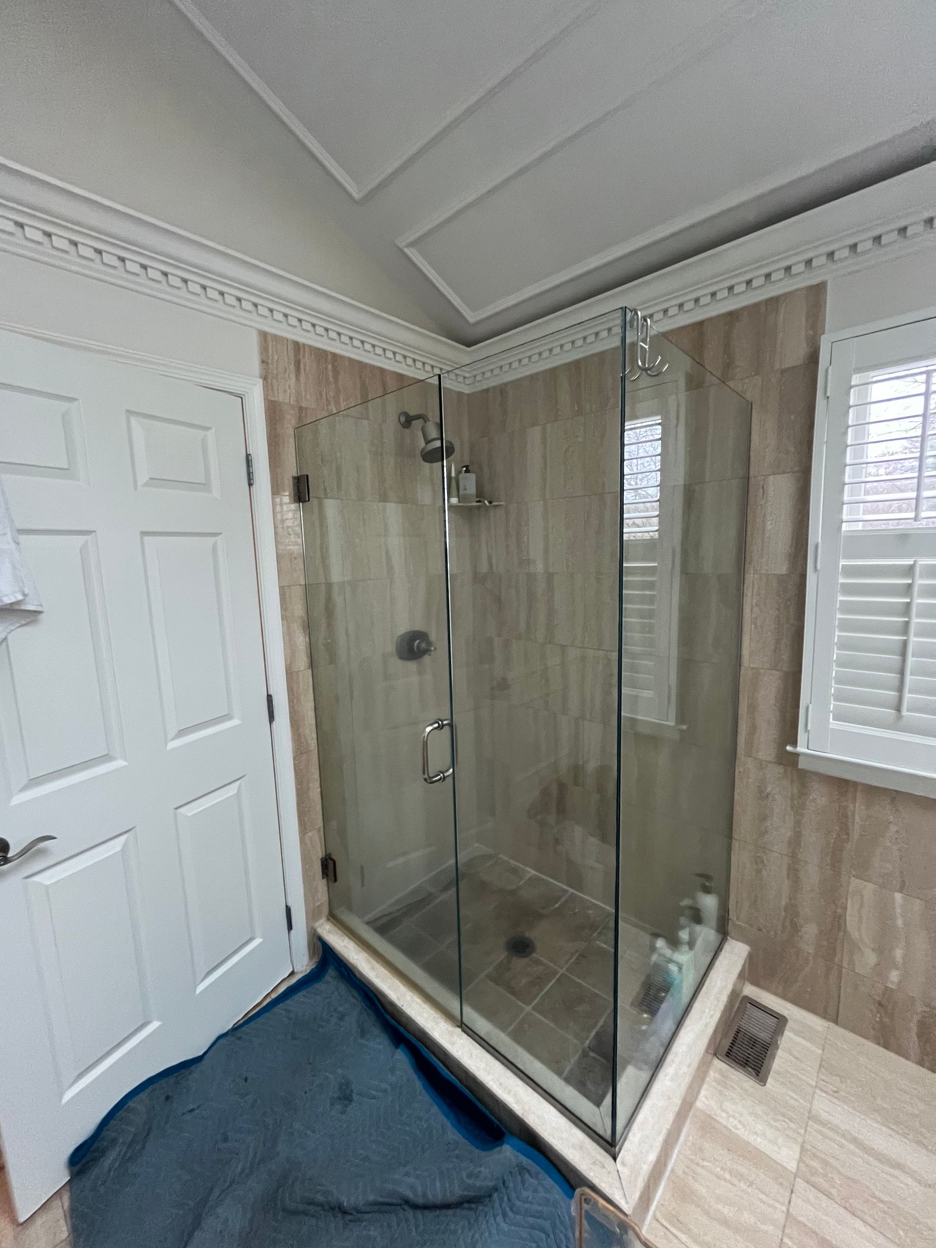 Corner glass shower stall in a bathroom with light-colored tile and a window with white shutters.