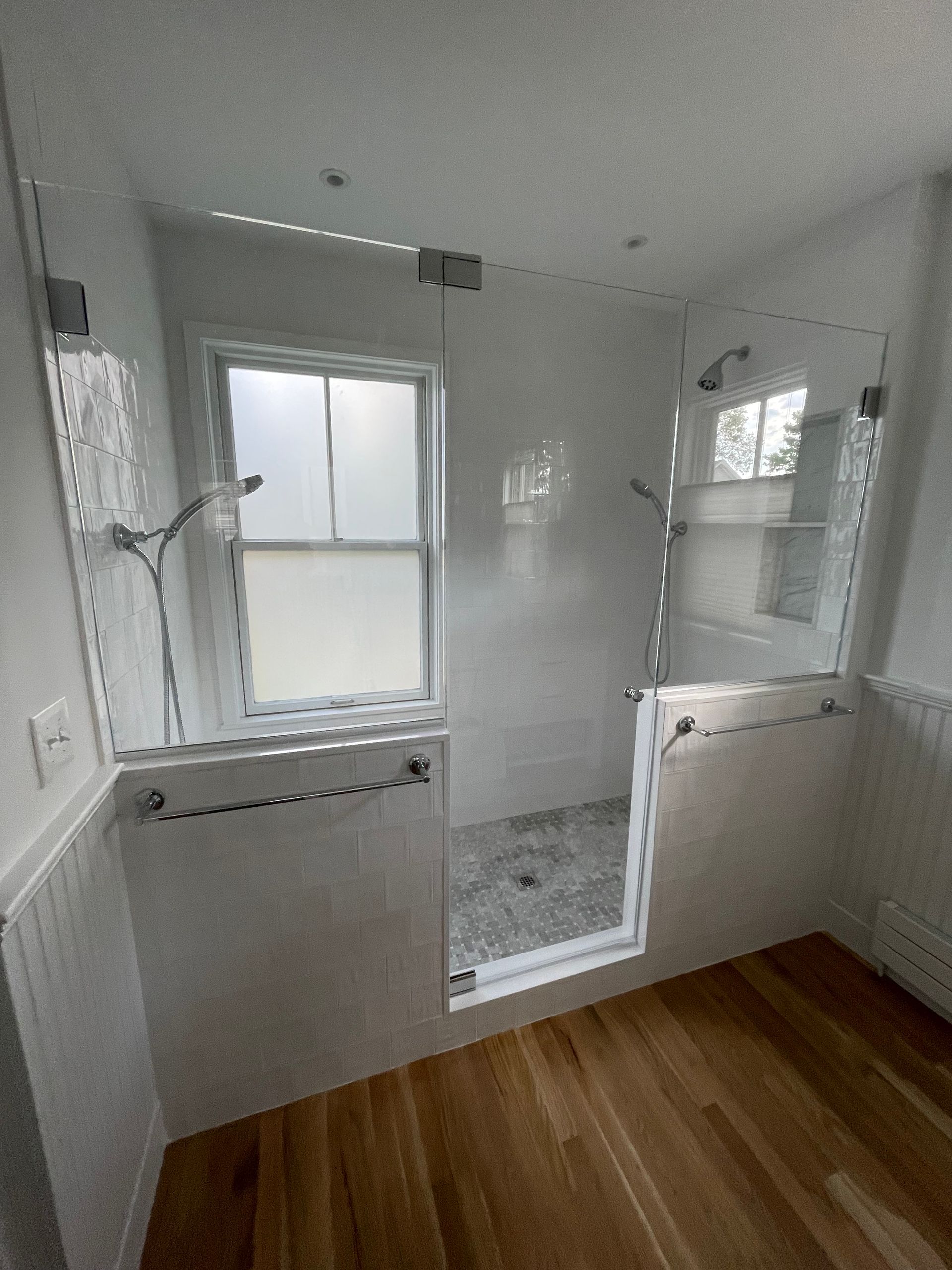 Bathroom with glass shower doors, white tile, window with frosted glass, and wood floor.