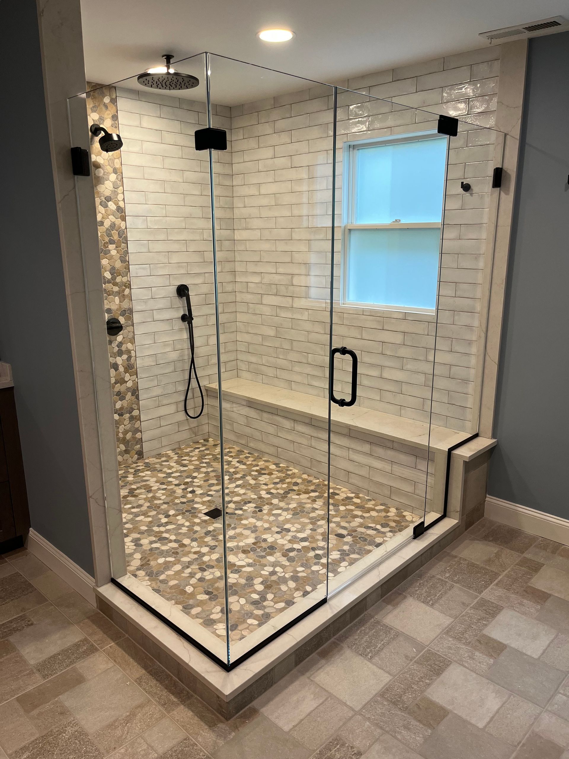 Modern shower with glass doors, stone bench, pebble floor, and white tile walls.