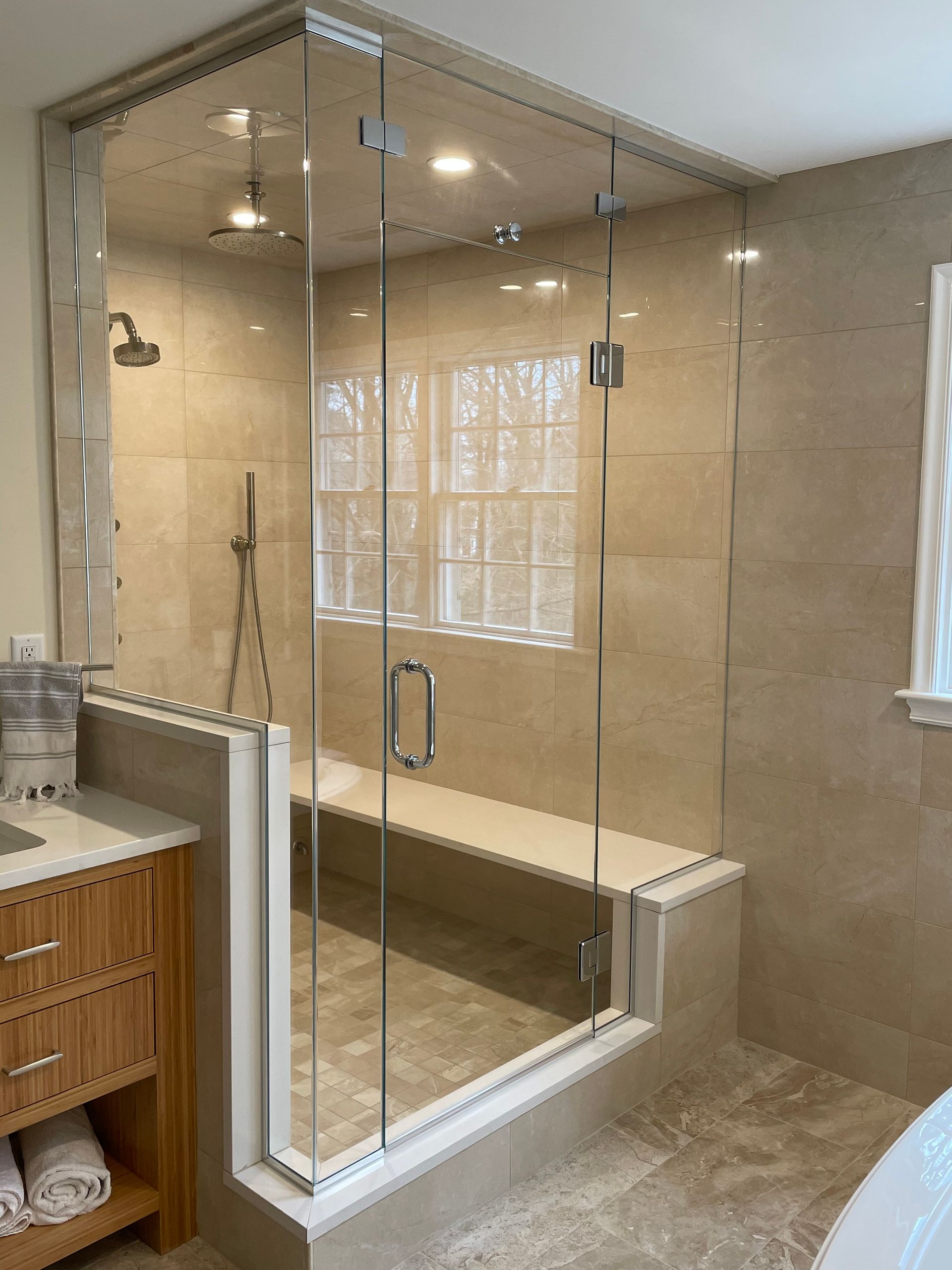 Glass-enclosed shower in a beige-tiled bathroom with a built-in bench. Natural light streams in from a window.