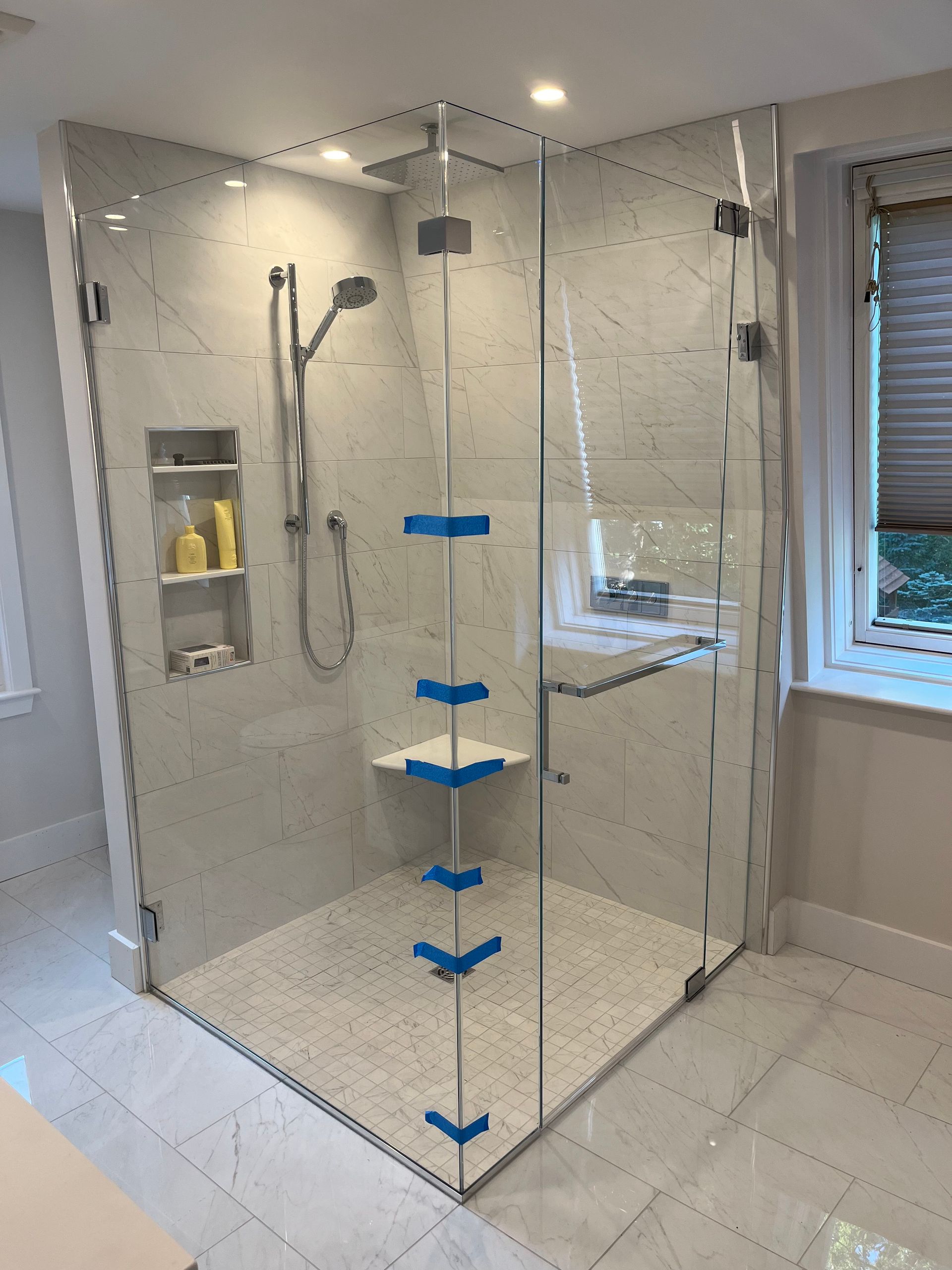 Glass shower enclosure with white tile walls and floor, corner seat, and blue tape markings.