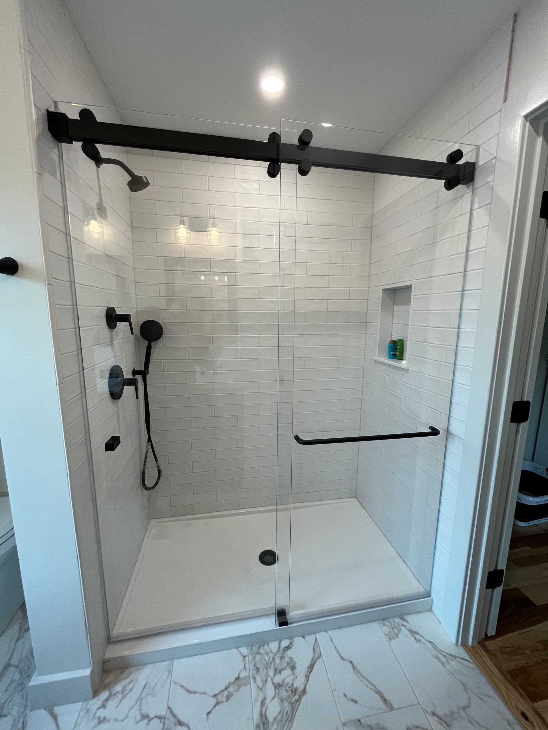 Modern shower with black fixtures and sliding glass doors. White tile walls and marble floor.