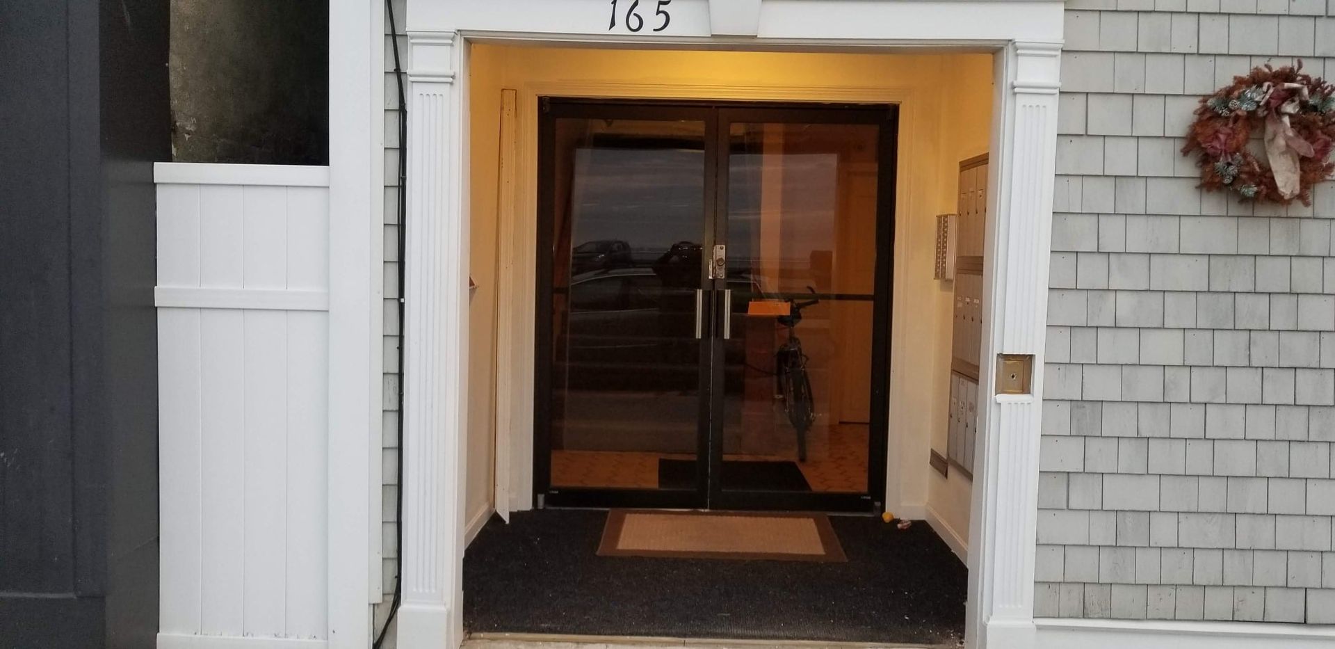 Front entrance with glass doors, a welcome mat, and a wreath. The building is gray, and the door frame is white.