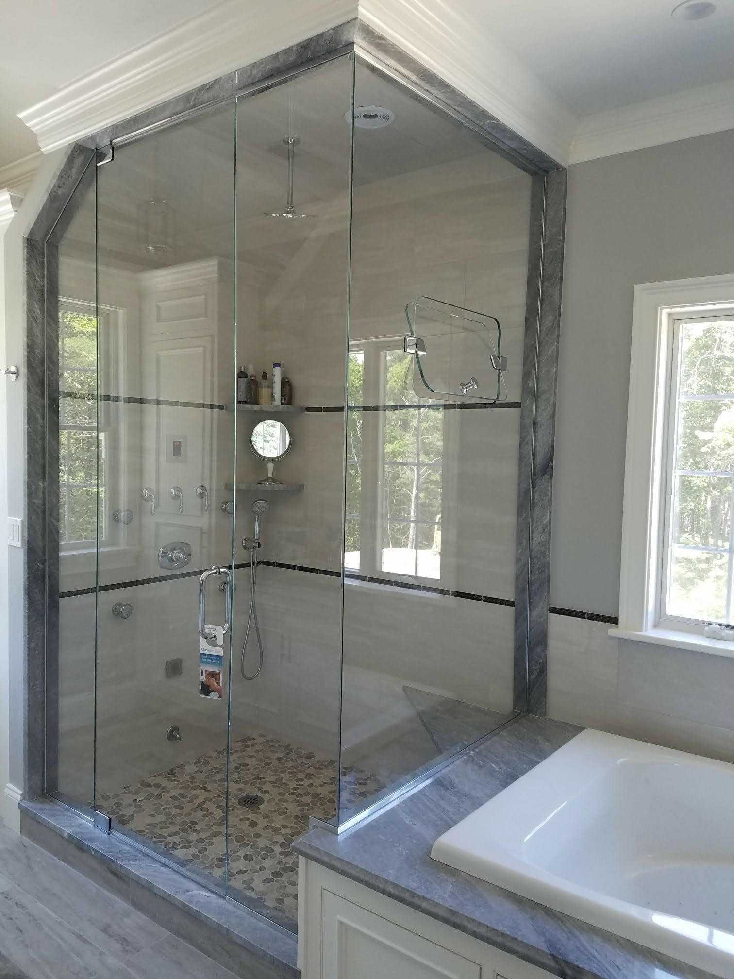A glass shower with gray marble trim and pebbled floor, beside a white tub in a bathroom.