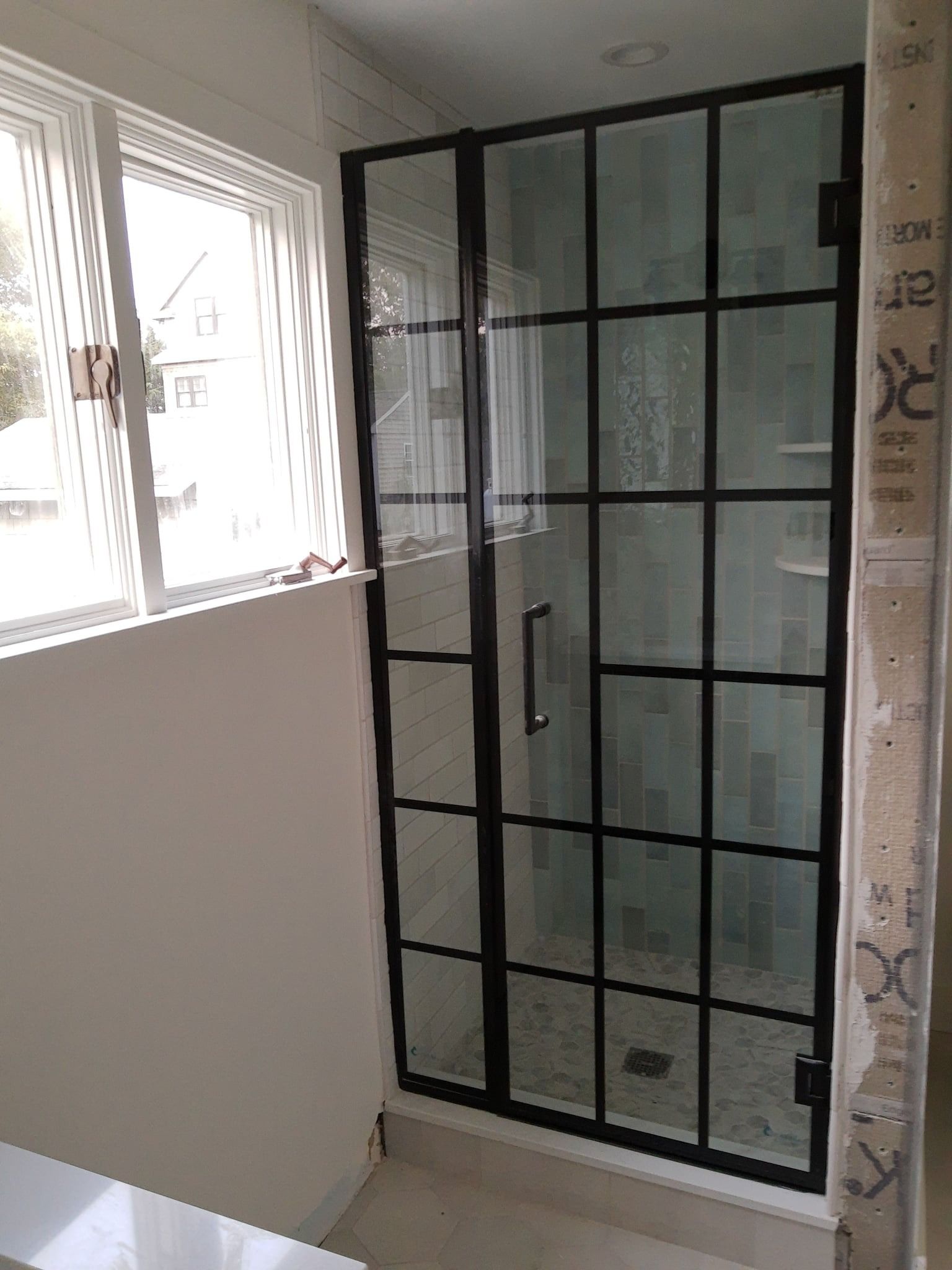 Black-framed shower door with grid design; adjacent to a window. White walls and neutral floor tiles are present.