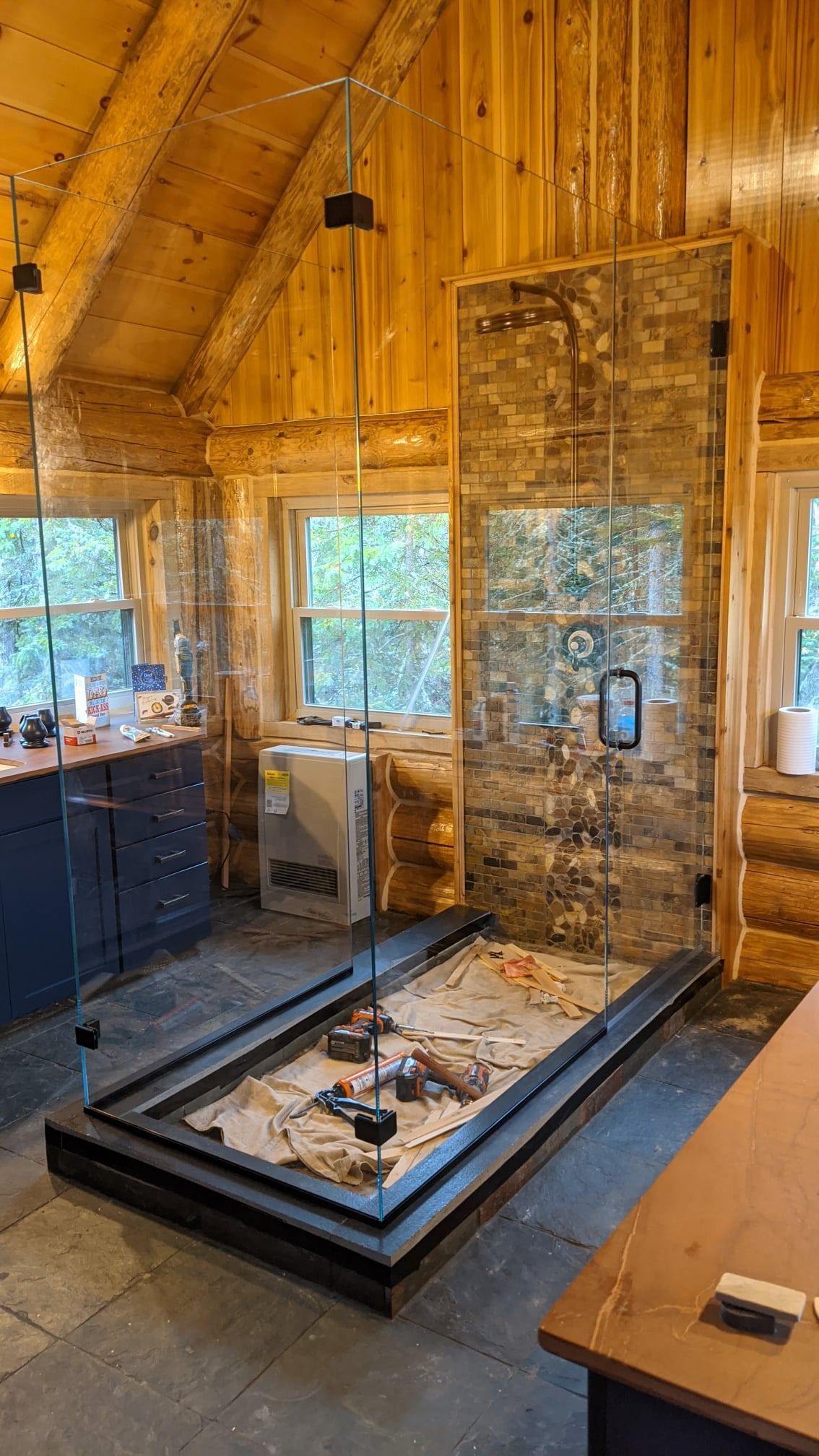 Glass-enclosed shower in a wood-paneled bathroom with a stone accent wall, on a raised platform.
