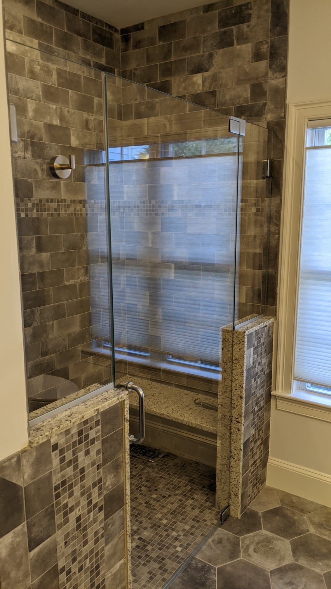 A tiled shower with a glass door, window with blinds in the back, and gray hexagon floor tiles.