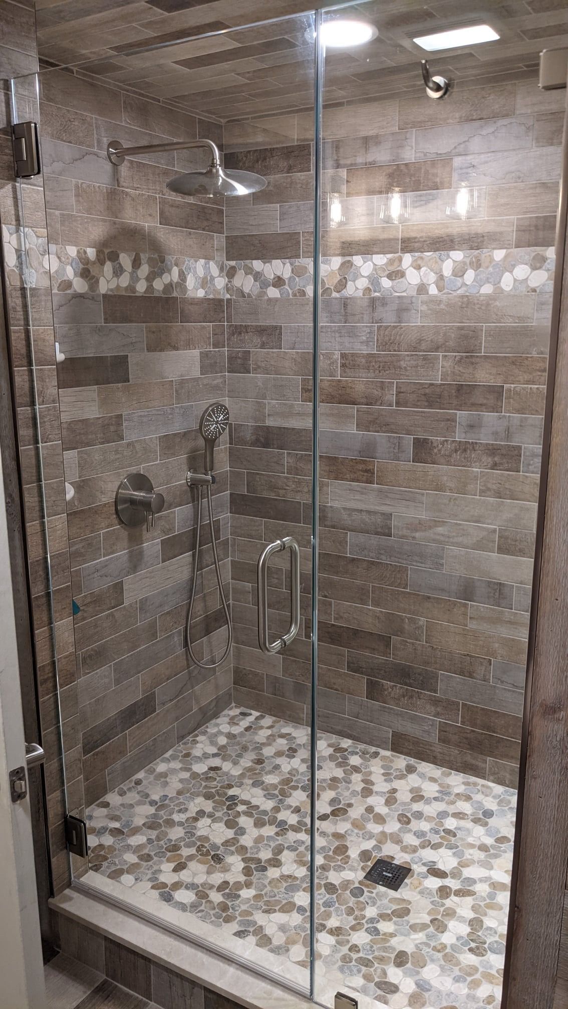 Glass-enclosed shower with brown, textured tile walls and pebble stone floor. Chrome fixtures.