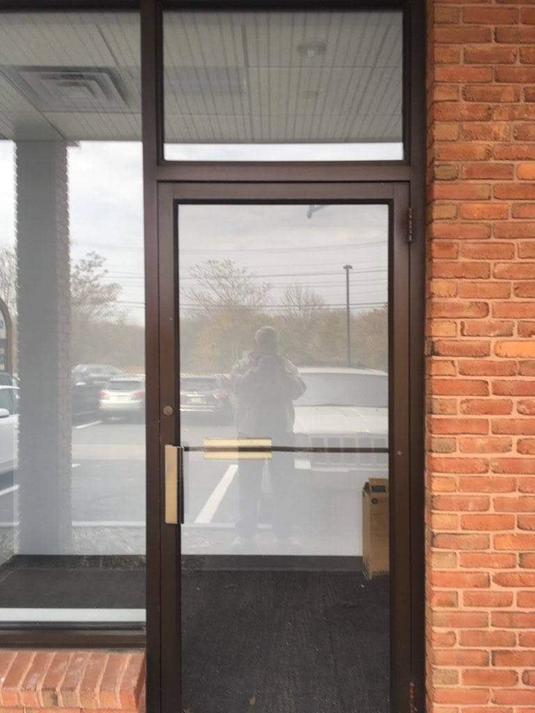 Brown-framed glass door with a push bar, next to a glass window. Brick wall on the right, cars visible outside.