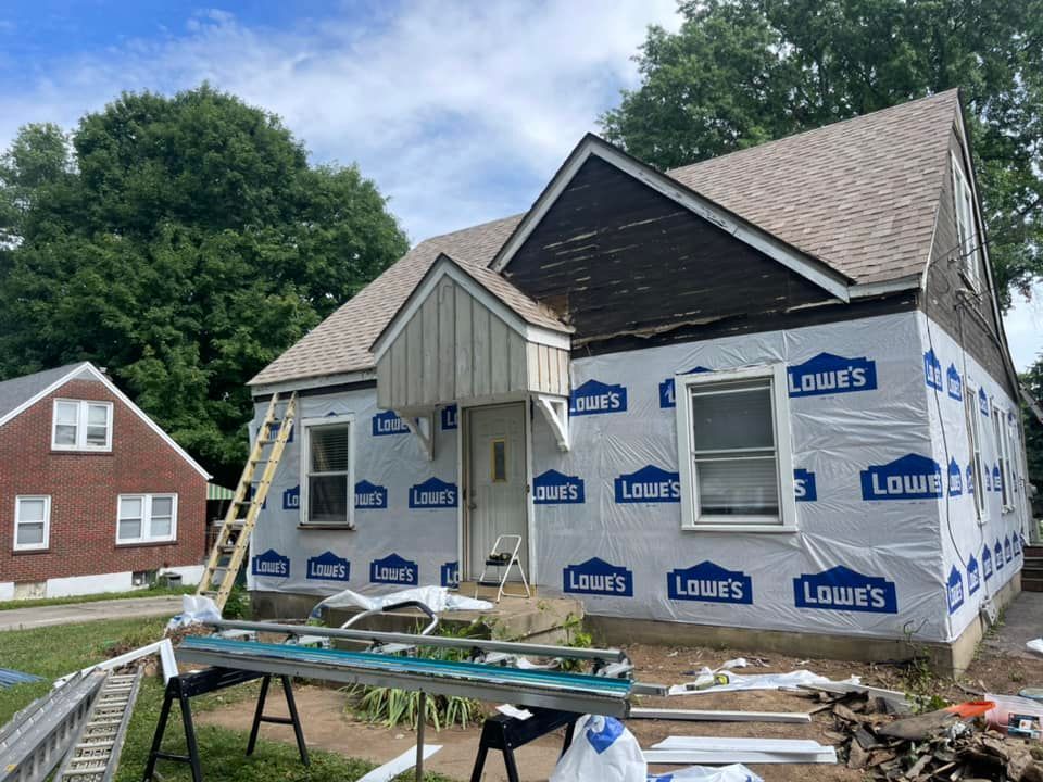 House exterior under renovation, covered with blue Lowe's wrap. A ladder is leaning against it.