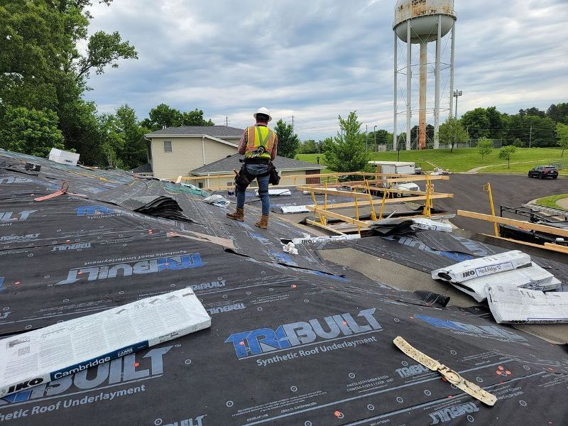 Roofer on a roof wearing a safety vest. Roof has black underlayment, building and water tower in background.