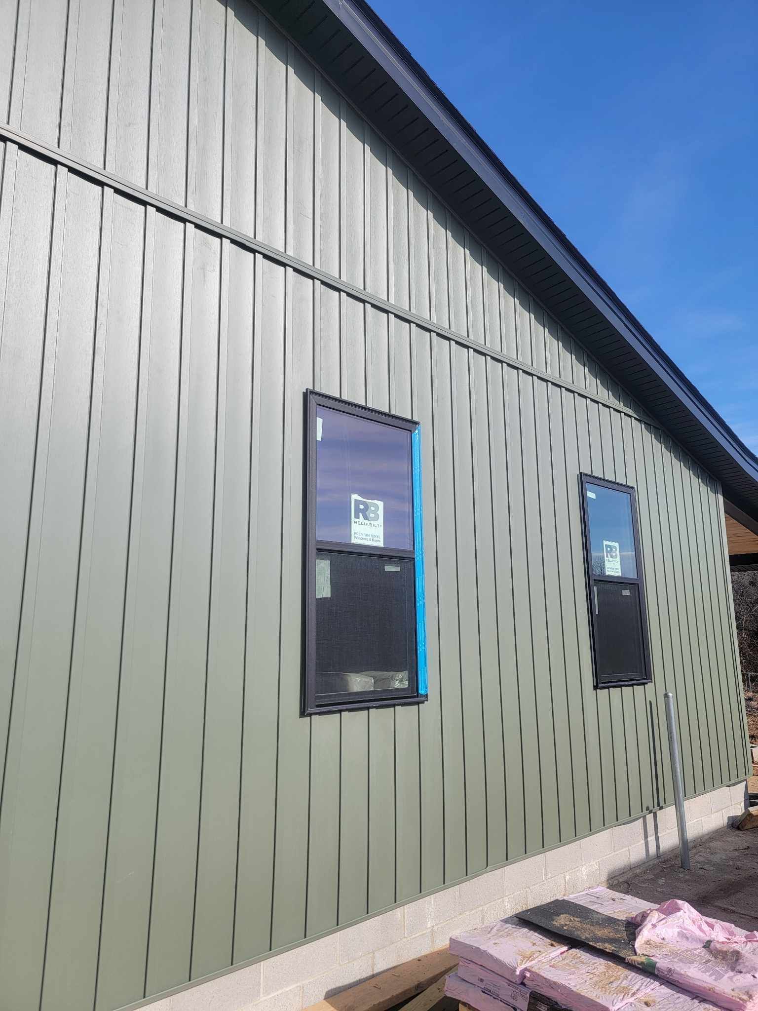 Green building exterior with black-framed windows. Construction materials on ground, under a clear, blue sky.