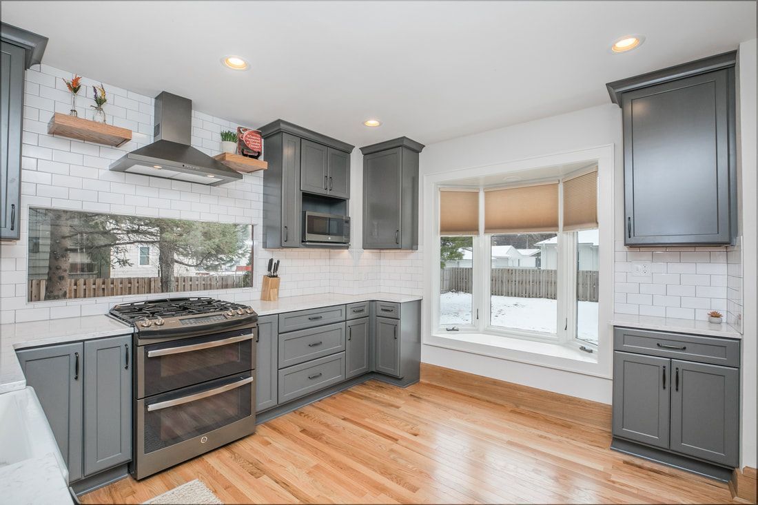 A kitchen with gray cabinets , stainless steel appliances , hardwood floors and a large window.
