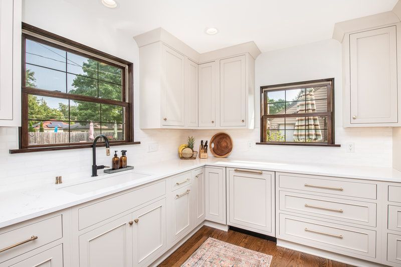 A kitchen with white cabinets , a sink , and two windows.