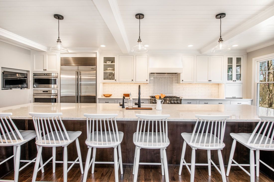 A kitchen with white cabinets , stainless steel appliances , a large island and chairs.