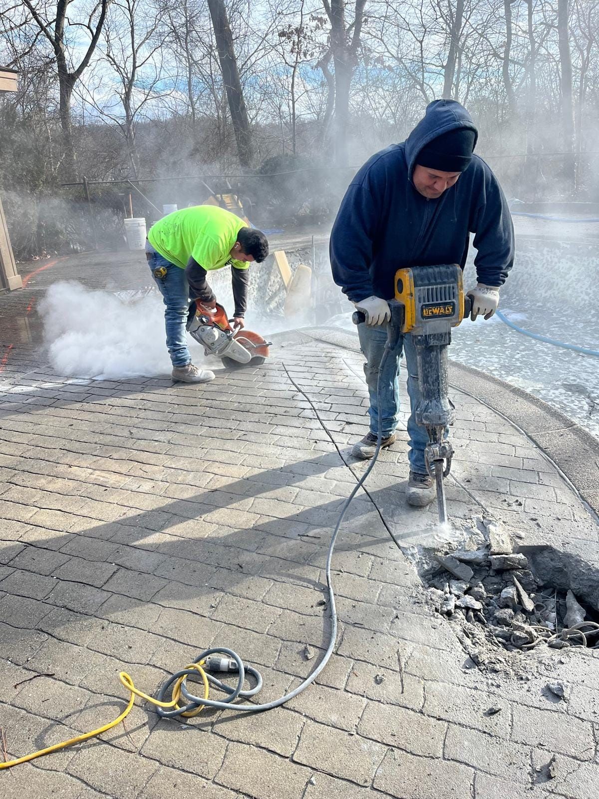 Two men are working on a patio with a hammer and a chainsaw.