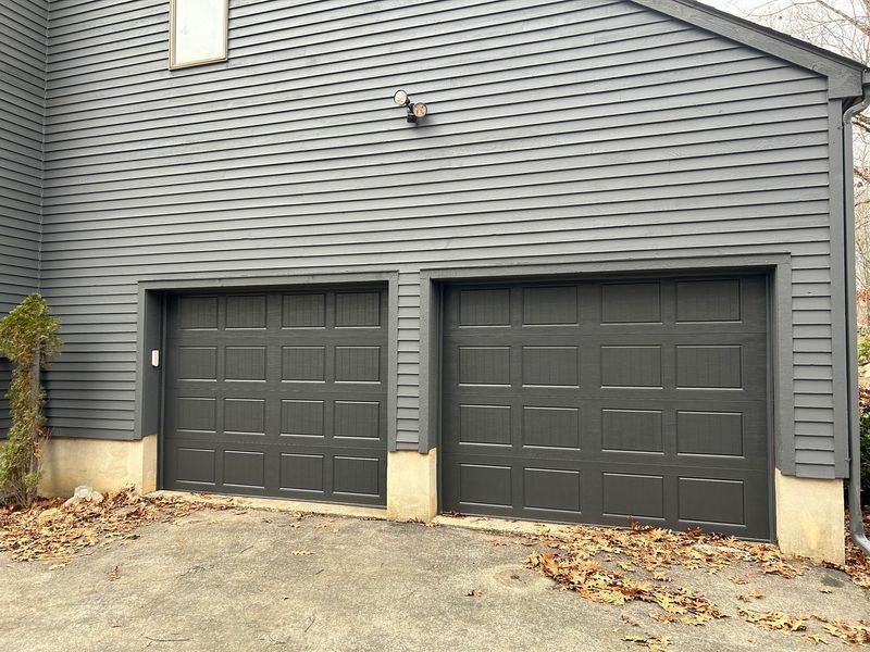Two-car gray garage with gray siding, two closed garage doors, tan trim, and leaves on the ground.