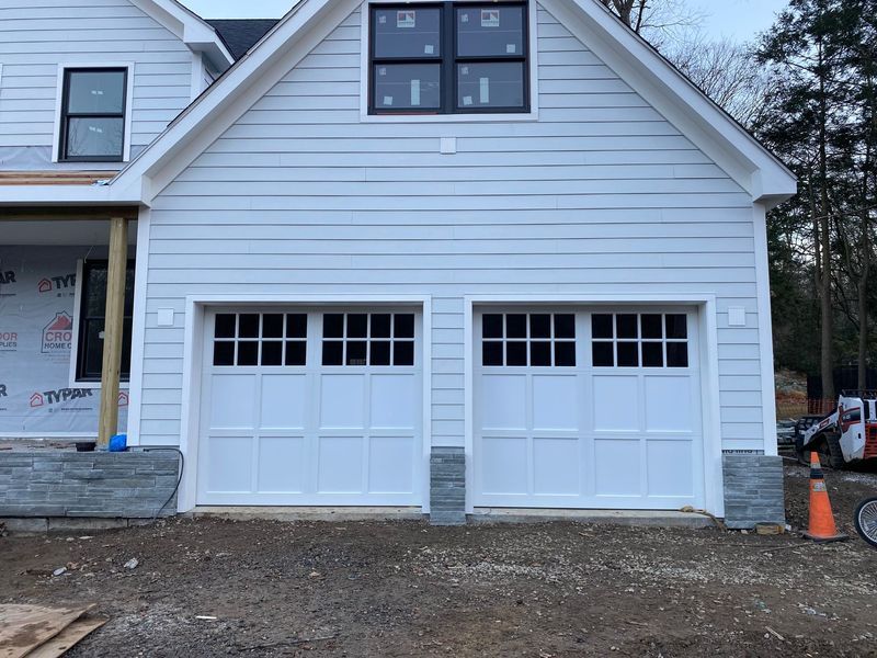 Two white garage doors with black-framed windows, light blue siding, and stone accents.