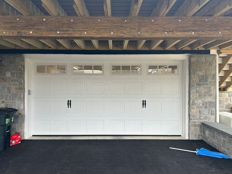 White garage door with windows, black handles, set under wooden beams and stone columns.