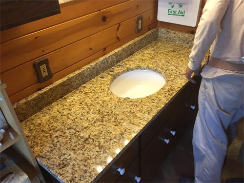 Granite countertop with oval sink, dark cabinets, and a person standing beside it in a wood-paneled bathroom.