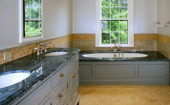 Bathroom with marble counters, grey cabinets, and a soaking tub under a window.