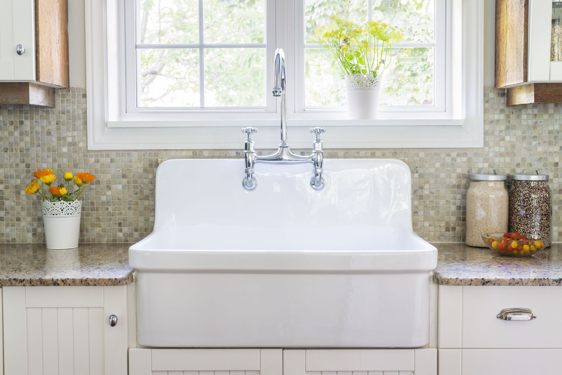 White farmhouse kitchen sink in front of a window, with flowers and glass jars on the counter.