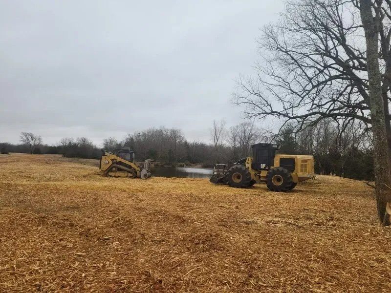 Two yellow construction machines clearing a field of golden-brown mulch, under an overcast sky.