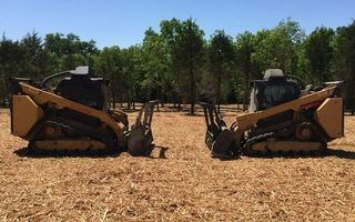 Two skid steer loaders facing each other, with tree shear attachments. Brown ground, trees in the background, sunny day.