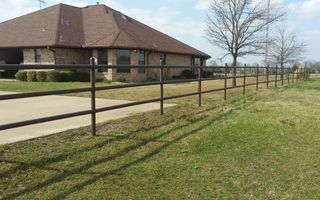 Brown metal fence lines a driveway next to a grassy lawn, with a brick house in the background.