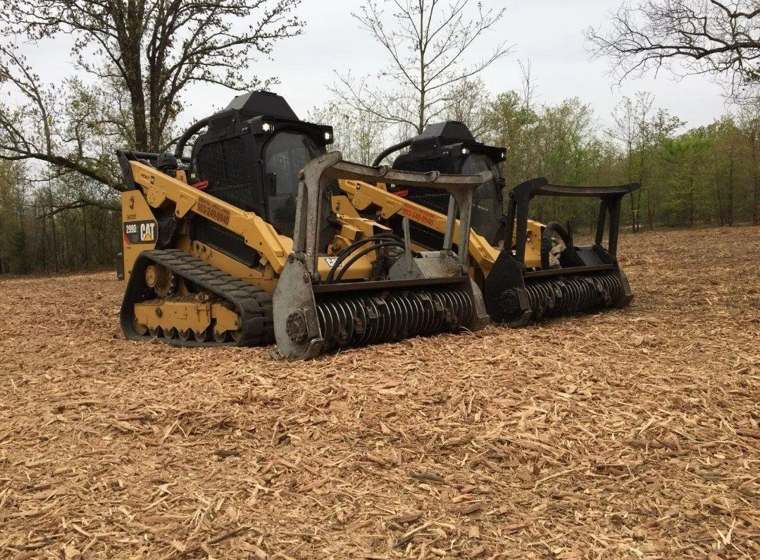 Yellow and black skid steer clearing debris in a field; trees in background.